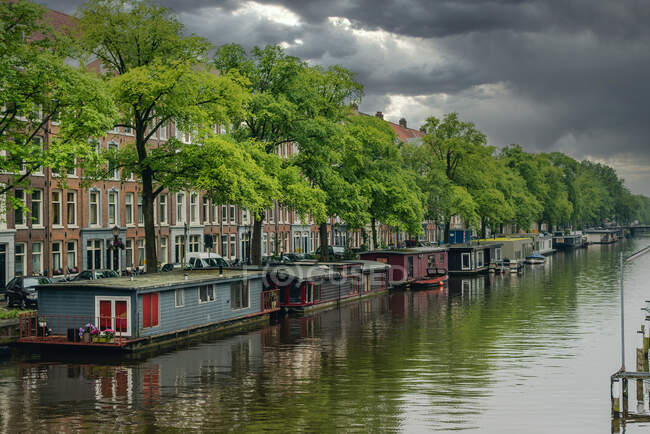 Aguas tranquilas del canal de la ciudad antes de la tormenta con nubes oscuras sobre edificios y árboles verdes en Amsterdam - foto de stock