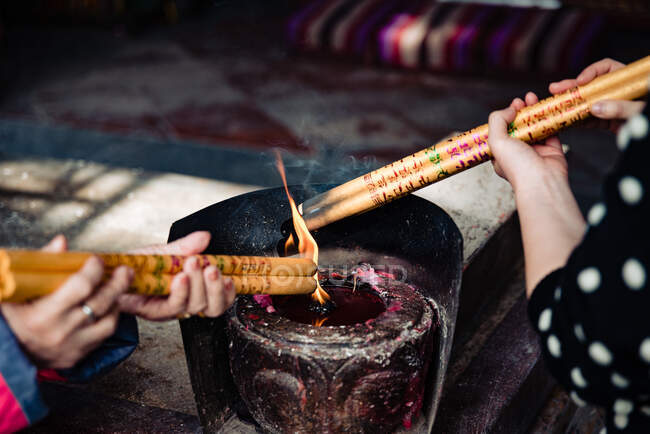 De cima de mulheres sem rosto segurando espessas varas de incenso chinês acima da luz com fumaça acima — Fotografia de Stock