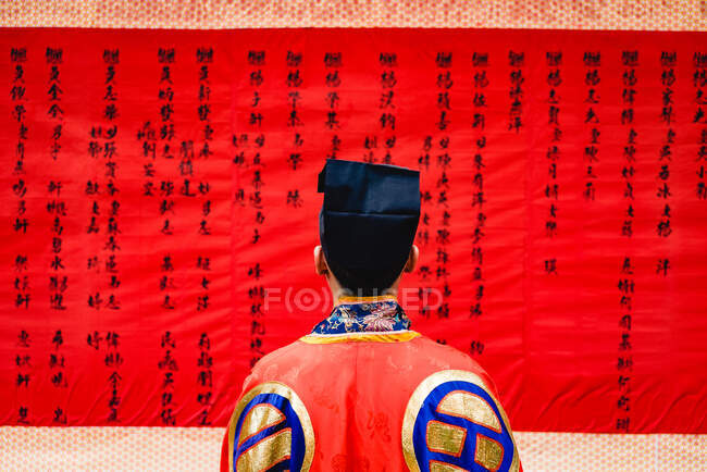 Visão traseira da pessoa anônima vestindo roupas chinesas coloridas olhando para o tecido vermelho com hieróglifos desenhados — Fotografia de Stock