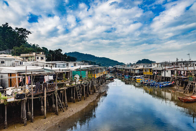 Vue en perspective d'une rivière paisible avec de petites maisons et des piliers en bois des deux côtés sous un ciel nuageux brillant à Hong Kong — Photo de stock