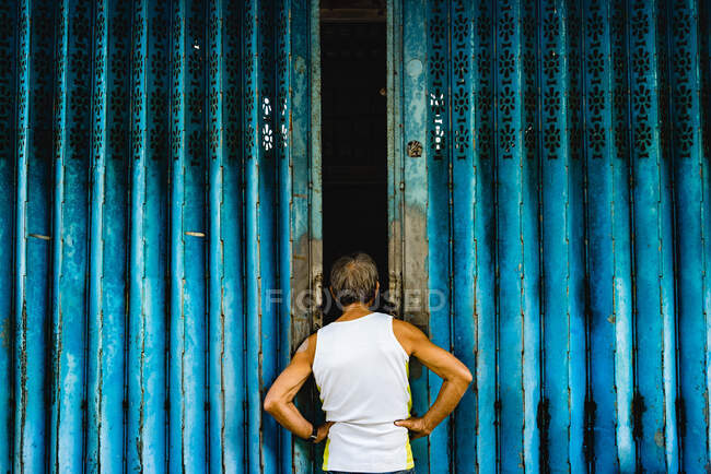 Back view of aged man in tank top holding arms on waist while looking inside of blue doorway in Hong Kong — Stock Photo
