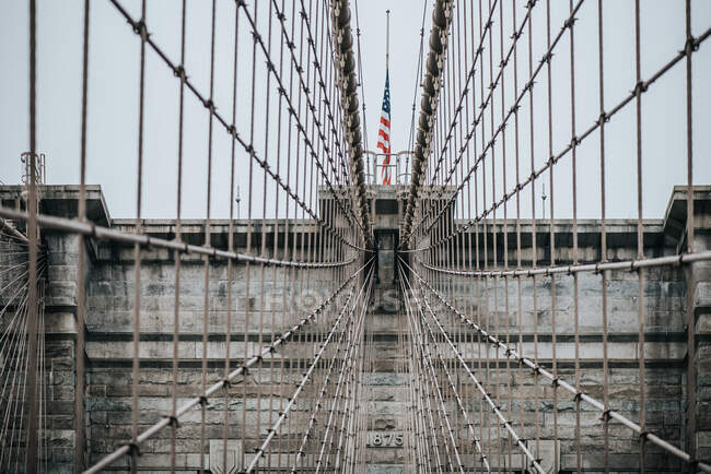 Bajo ángulo de torres de piedra con arcos puntiagudos de puente colgante de Brooklyn con bandera estadounidense en día nublado - foto de stock