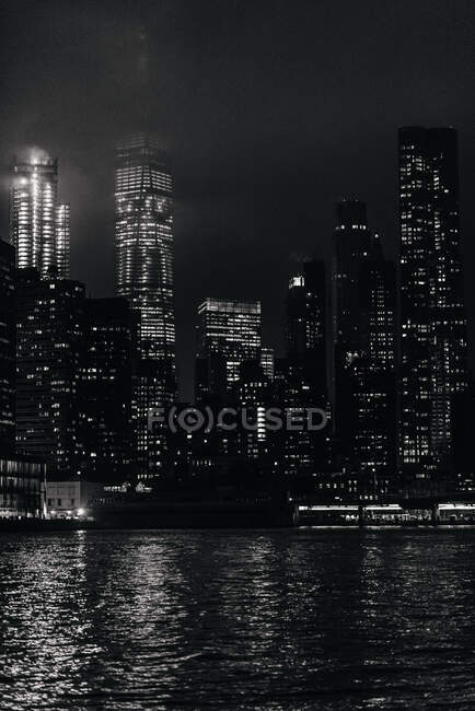 Complesso in bianco e nero di moderni skyline illuminati a Manhattan situato di fronte a un fiume calmo durante la notte — Foto stock