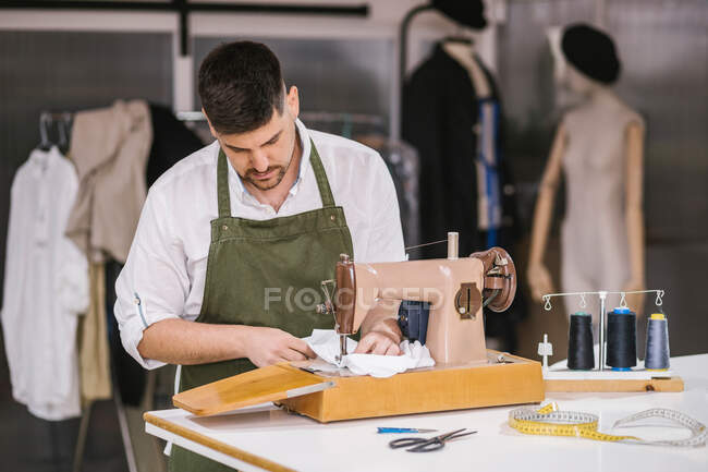 Diligent focused male tailor in apron sewing outfit details using modern sewing machine at table while creating exclusive clothes collection in contemporary work studio — Stock Photo