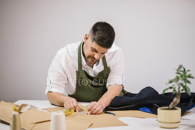 Costurera masculina usando cinta métrica mientras comprueba el tamaño exacto de los patrones mientras hace atuendo a medida para el cliente en el taller moderno - foto de stock