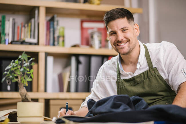 Crop happy young male dressmaker working developing details of future outfit while working on exclusive apparel collection at table against blurred interior of contemporary atelier — Stock Photo