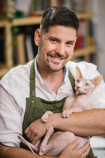 Alegre artesano sin afeitar en camisa blanca y delantal verde sonriendo a la cámara mientras lleva tranquilo gato Esfinge en las manos en el estudio moderno - foto de stock