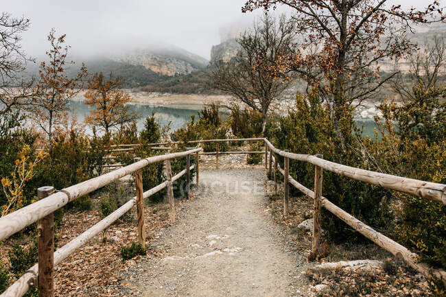 Caminho curvo vazio com trilhos de madeira que levam entre árvores com folhagem seca na encosta da colina perto do lago da montanha em dia nebuloso na cordilheira Montsec, na Espanha — Fotografia de Stock