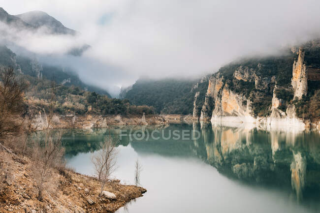 Magnífica paisagem de lago calmo com superfície de água espelhada cercada por montanhas rochosas ásperas de Montsec Range coberto com densa névoa em dia frio na Espanha — Fotografia de Stock