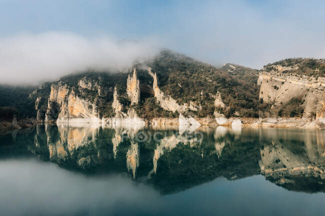 Чудовий краєвид спокійного озера з дзеркальною поверхнею води в оточенні скелястих гір хребта Монсек, вкритих густим туманом в холодні дні дні дні в Іспанії. — стокове фото