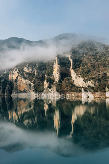 Magnífico paisaje de lago tranquilo con superficie de agua espejada rodeada de ásperas montañas rocosas de Montsec Cordillera cubierta de densa niebla en día frío en España - foto de stock