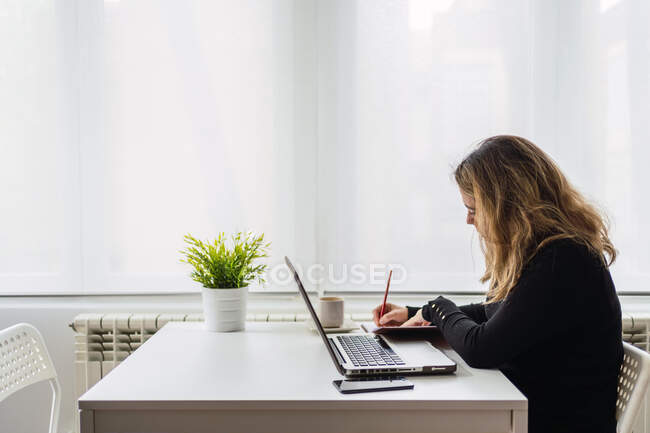 Vista lateral de la joven mujer enfocada en traje casual sentada en la mesa con el ordenador portátil y tomando notas en el planificador mientras trabaja en línea en el lugar de trabajo ligero en casa - foto de stock