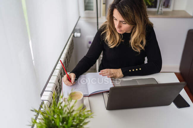 From above of focused young female in casual outfit sitting at table with laptop and taking notes in planner while working online in light workplace at home — Stock Photo