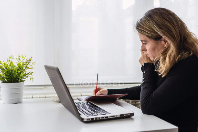 Вид сбоку сосредоточенной молодой женщины в повседневной одежде, сидящей за столом с ноутбуком и делающей заметки в планировщике, работая дома на легком рабочем месте — стоковое фото