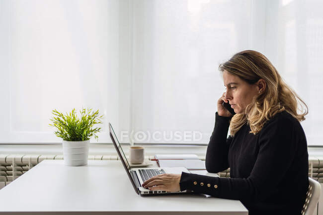 Vista lateral de la joven mujer enfocada en atuendo casual sentada en la mesa con computadora portátil y teléfono hablando mientras trabaja en línea en el lugar de trabajo ligero en casa - foto de stock