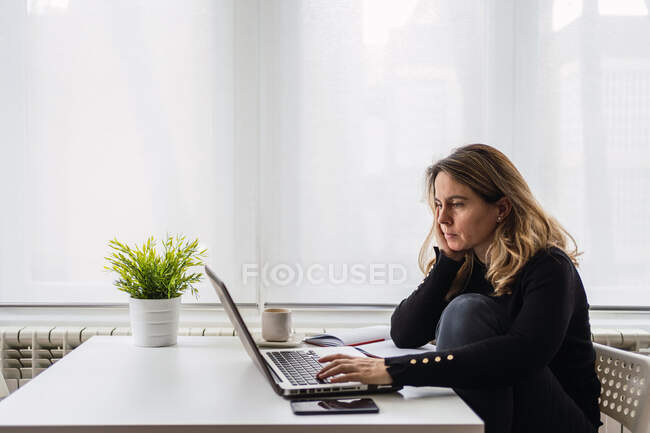 Vista lateral de ángulo alto del especialista femenino enfocado en ropa casual que trabaja con documentos electrónicos en el ordenador portátil mientras está sentado en la mesa cerca de la ventana en la habitación moderna en casa - foto de stock