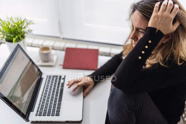 Високий кут зору зосередженої жінки-спеціалістки в повсякденному носінні, що працює з електронними документами на ноутбуці, сидячи за столом біля вікна в сучасній кімнаті вдома — стокове фото