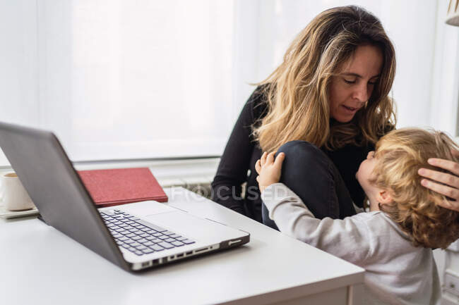 Современная занятая предпринимательница в повседневной одежде обнимает и целует малыша во время работы над проектом с ноутбуком в домашнем офисе — стоковое фото