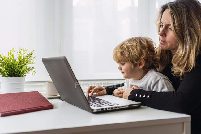 Side view of female remote employee holding curious little child on knees while sitting at table and working with laptop at home — Stock Photo
