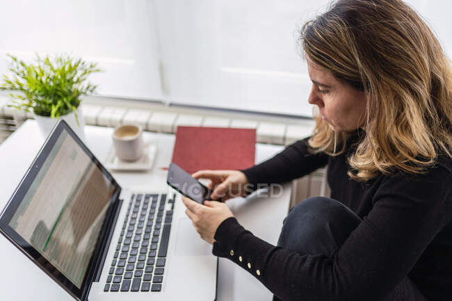 Зверху зосереджена жінка-спеціалістка в повсякденному одязі, що працює з електронними документами на ноутбуці, сидячи за столом біля вікна в сучасній кімнаті вдома — стокове фото