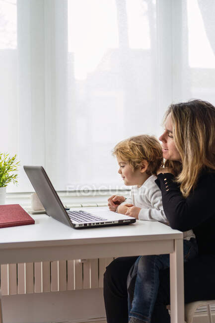 Vista lateral de la empleada remota femenina sosteniendo a un niño pequeño y curioso de rodillas mientras está sentado en la mesa y trabajando con el portátil en casa - foto de stock