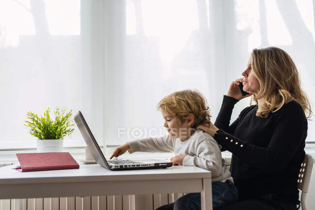 Vista lateral da funcionária remota feminina segurando uma criança curiosa de joelhos enquanto se senta à mesa e trabalha com laptop em casa — Fotografia de Stock