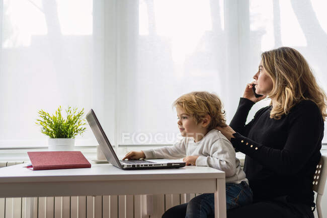 Vista laterale di un'impiegata remota che tiene in ginocchio un bambino curioso mentre si siede a tavola e lavora con il computer portatile a casa — Foto stock
