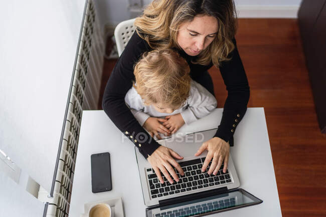 De cima de colheita funcionário remoto feminino segurando pequena criança curiosa de joelhos enquanto sentado à mesa e trabalhando com laptop em casa — Fotografia de Stock