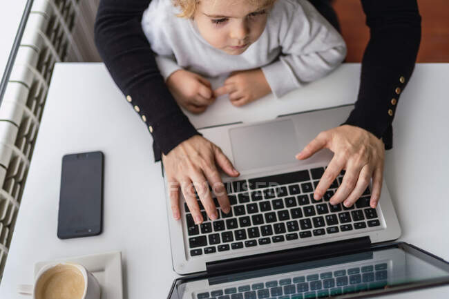 De cima de colheita anônima funcionário remoto feminino segurando pequena criança curiosa de joelhos enquanto sentado à mesa e trabalhando com laptop em casa — Fotografia de Stock
