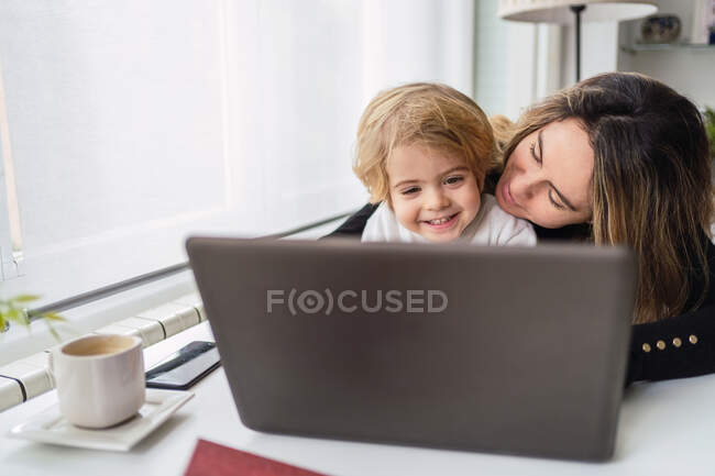 Female remote employee holding curious little child on knees while sitting at table and working with laptop at home — Stock Photo