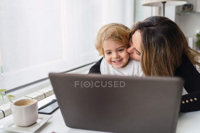 Empleada remota que sostiene a un niño pequeño y curioso de rodillas mientras está sentada en la mesa y trabaja con la computadora portátil en casa - foto de stock