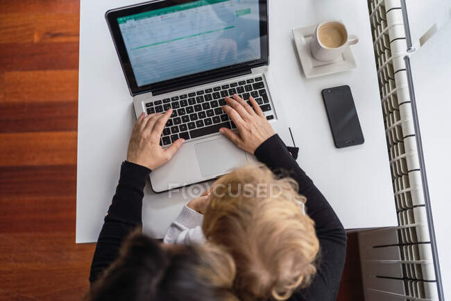 From above of crop anonymous female remote employee holding curious little child on knees while sitting at table and working with laptop at home — Stock Photo