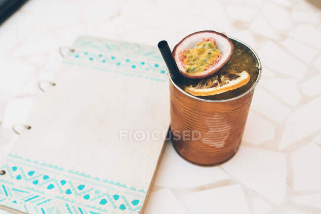 Delicioso cocktail refrescante com maracujá servido em lata de metal com palha colocada perto da página de papel com espaço em branco na mesa — Fotografia de Stock