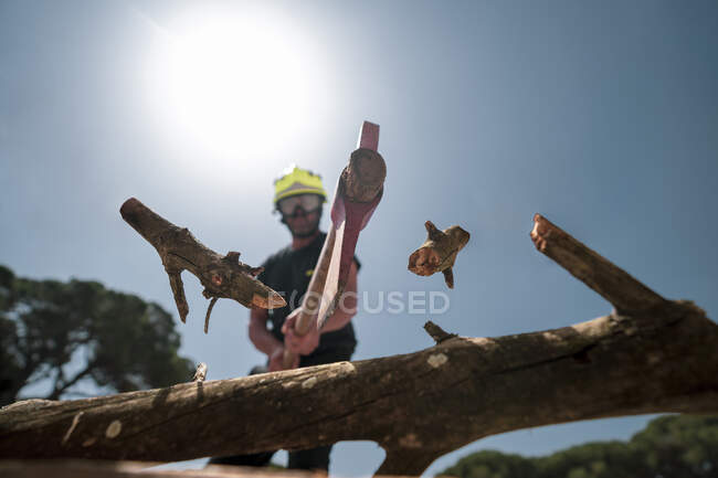 From below of fireman in protective uniform cutting branch with ax in wood on background of blue sky — Stock Photo