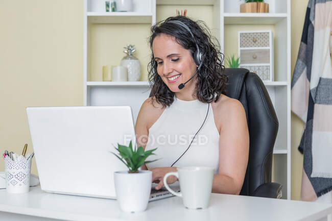 Усмішка в білій блузці сидить за столом з чашкою гарячого напою і використовує навушники, працюючи вдома на ноутбуці. — стокове фото