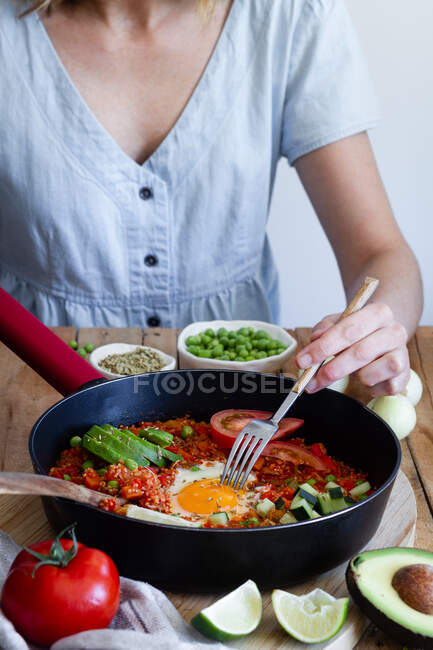 Невизначена жінка в одязі, сидячи за столом з різними інгредієнтами і насолоджуючись свіжою шейшукою з яйцями та овочами — стокове фото