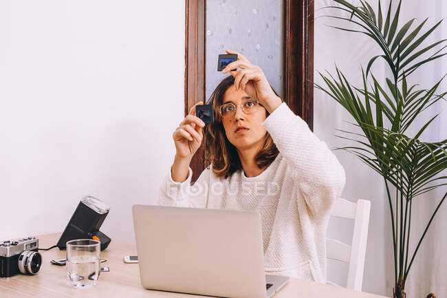 Giovane fotografa donna seduta a tavola con computer portatile e macchina fotografica con proiettore e che lavora con vecchie diapositive fotografiche sul posto di lavoro di casa — Foto stock