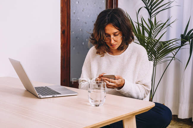 Seitenansicht einer fokussierten jungen Freiberuflerin in lässiger Kleidung, die Nachrichten auf dem Smartphone checkt, während sie mit Laptop am Tisch sitzt und zu Hause an einem Projekt arbeitet — Stockfoto