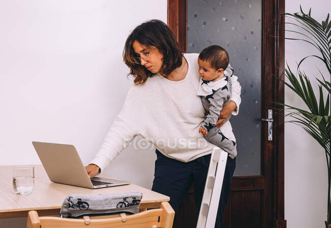 Молода зайнята жінка в повсякденному одязі з маленькою дитиною під рукою стоїть за столом і перевіряє електронну пошту на ноутбуці під час роботи онлайн вдома — стокове фото