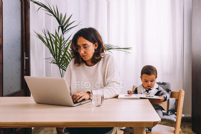 Jeune femme pigiste en vêtements décontractés assis à la table et travaillant sur le projet avec ordinateur portable tandis que le petit enfant assis à proximité avec livre — Photo de stock