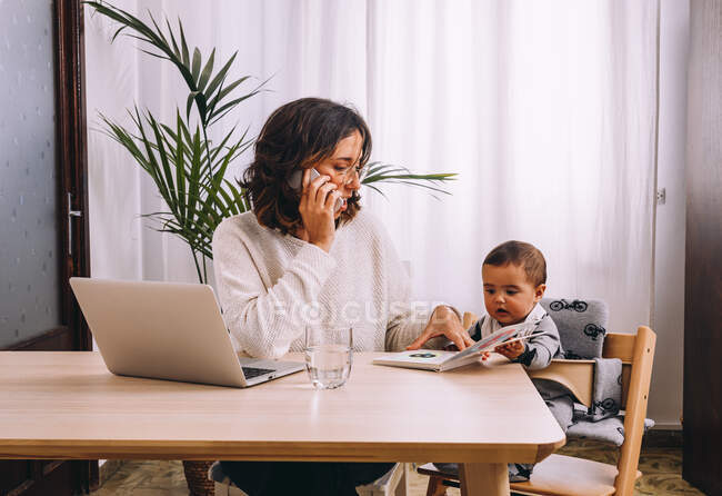 Jeune femme pigiste en vêtements décontractés assise à table et travaillant sur un projet avec ordinateur portable et parlant sur le téléphone portable tandis que le petit enfant assis à proximité avec livre — Photo de stock