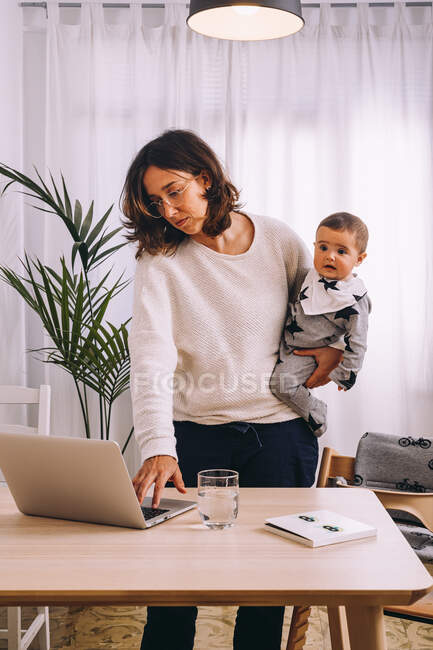Joven mujer ocupada en ropa casual con un niño pequeño en la mano de pie en la mesa y comprobando el correo electrónico en el ordenador portátil mientras trabaja en línea en casa - foto de stock