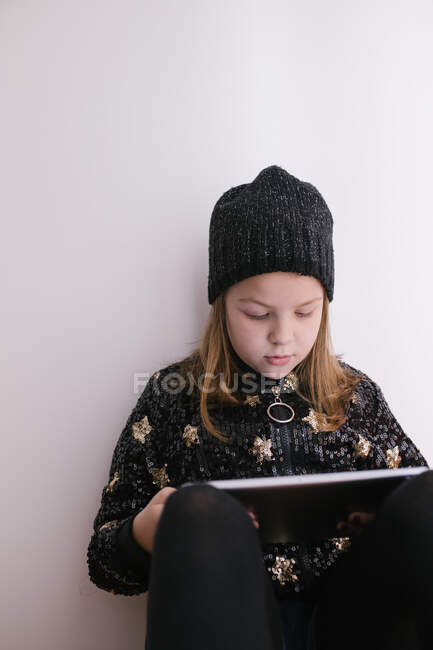 Chica pensativa en suéter cálido y gorra de punto navegar por Internet en la tableta mientras está sentado apoyado en la pared blanca - foto de stock