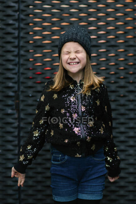 Cheerful child with toothy smile in warm black jersey and knitted cap covering eyes while standing on green background — Stock Photo
