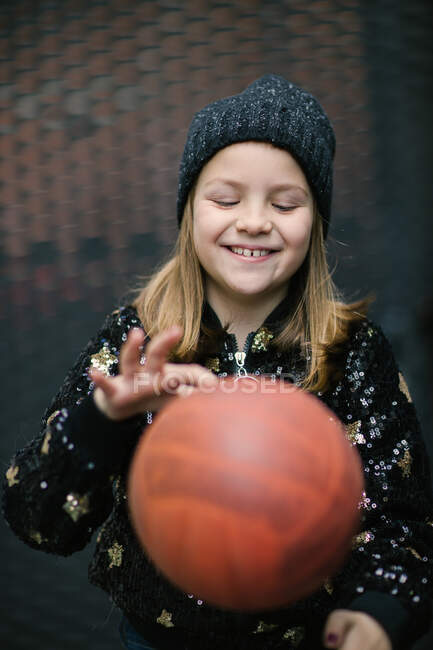 Niño feliz con espacio entre los dientes en gorra de punto y jersey caliente jugando con voleibol naranja al aire libre - foto de stock