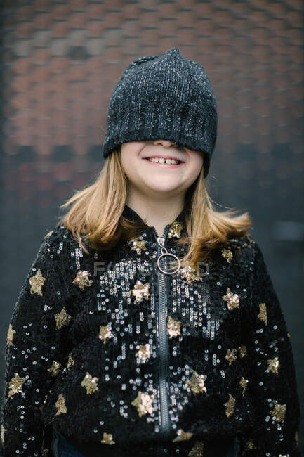 Divertido niño anónimo en chaqueta caliente y gorra de punto que cubre la mitad de la cara de pie al aire libre y sonriendo - foto de stock