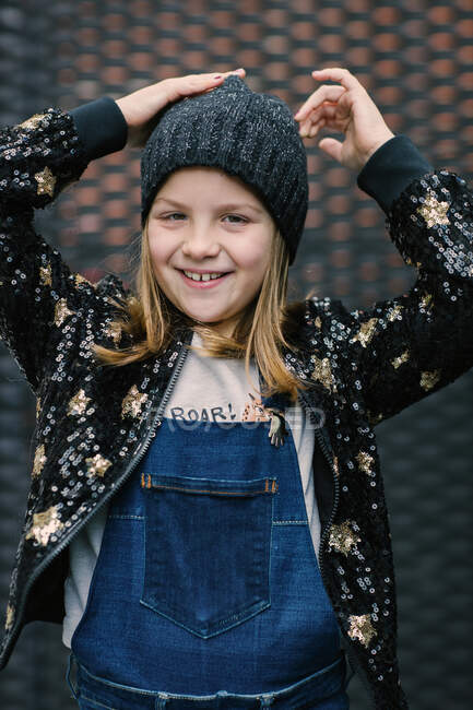 Enfant joyeux avec sourire de dents en jersey noir chaud et bonnet tricoté regardant la caméra tout en se tenant debout sur fond rouge — Photo de stock