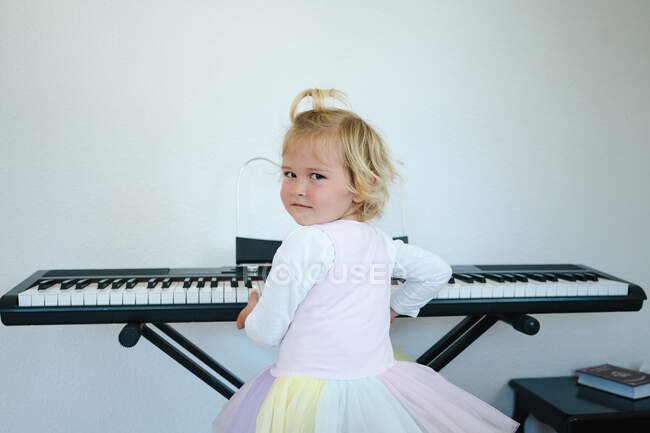 Back view of schoolgirl in fluffy skirt sitting at synthesizer and preparing for music class looking at camera — Stock Photo