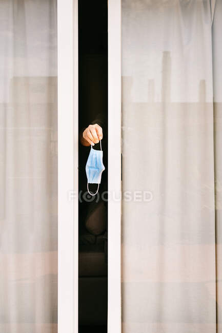 Mano de una persona anónima que sobresale de una ventana ligeramente abierta sosteniendo una máscara protectora que permanece en cuarentena durante el brote de coronavirus - foto de stock