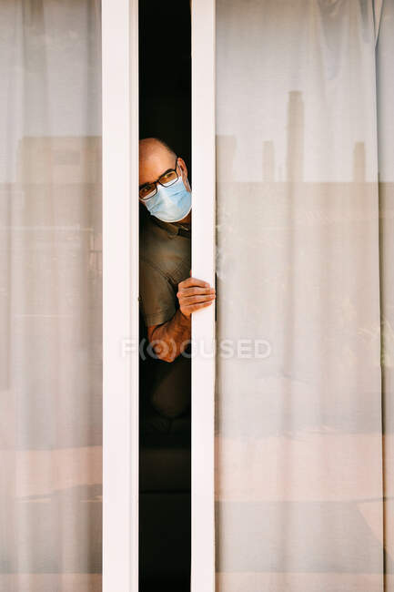 Adult female in medical mask looking out on street with fear during pandemic of COVID 19 — Stock Photo